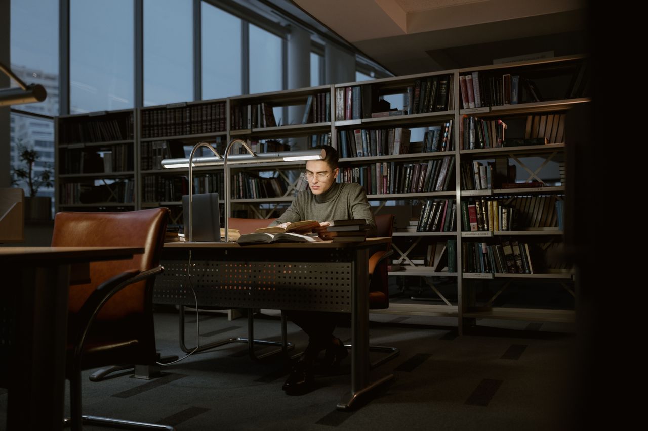A man intensely studies in a quiet library, surrounded by books and soft lighting.